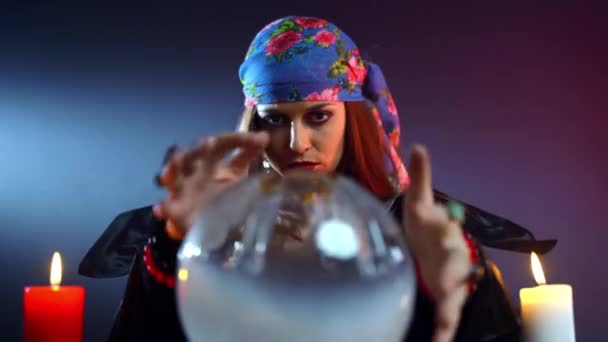 Frightful witch using round crystal ball for spelling over it, gypsy sitting in dark room and moving hands above crystal sphere surrounded with candles. Talking with black evil and afterlife — Stock Video