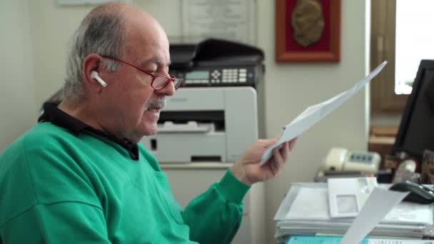 部屋に座っている老人は、ワイヤレスブルートゥースヘッドフォンを使用して、従業員と話をします。自宅の書類、検疫期間中の書類との長距離勤務の上級教授 — ストック動画