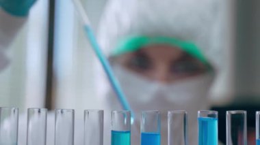 Koruyucu kıyafetli profesyonel kadın virolog mavi reaktör ve kimyasal elementlerle ilgili araştırma laboratuvarında önemli deneyler yapıyor. Kimyager steril test tüpleri ve şeffaf pipet kullanıyor.