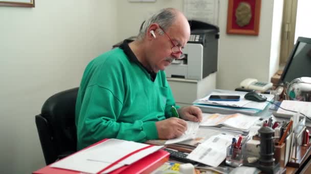 Trabajando a distancia en cuarentena de coronavirus, anciano con auriculares blancos inalámbricos sentado en el escritorio de su habitación y escribiendo algo con pluma, hombre mayor llenando facturas, leyendo — Vídeo de stock