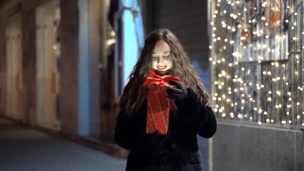 Mujer bastante sonriente sosteniendo regalo de Navidad con cinta roja en las manos, hermosa hembra en ropa de invierno abriendo misterio presente de pie en la calle sobre increíbles guirnaldas brillantes fondo. Mujer. — Vídeo de stock