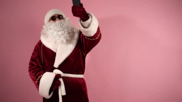 Кумедний Санта Клаус танцює на ізольованому рожевому фоні, людина робить онлайн-дзвінок на смартфоні, розмовляє з аудиторією в Інтернеті і посилає побажання. Santa Claus використовує мобільний телефон для онлайн — стокове відео