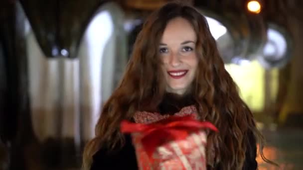 Retrato de mujer adorable con labios rojos mirando a cámara, bailando y sosteniendo regalo de Navidad en las manos. Mujer disfrutando de vacaciones de Navidad al aire libre, fuente de mármol iluminado con agua corriente en — Vídeo de stock