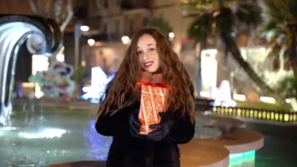 Vacker glad kvinna som håller speciella julklapp i händerna och dansa på fontän bakgrund och gata belysning på natten, leende kvinna njuter av festlig vinterkväll och visar henne verkliga — Stockvideo