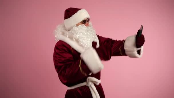 Trendy Santa Grandfather doing online video call on smartphone, talking with online audience and sending christmas wishes. Santa Claus browsing internet, christmas man dancing on isolated pink — Stock video