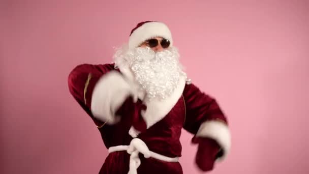 Claus santa insouciant en costume traditionnel rouge, avec des lunettes sombres et barbe bouclée dansant sur fond rose isolé et gesticulant avec les mains sur la caméra. drôle Santa profiter de la musique et de détente — Video