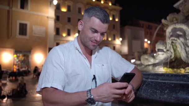 Positive video content creator using smartphone apps and chatting online with audience, young handsome man standing on illuminated street background with amazing ancient building exterior and marble — Stockvideo