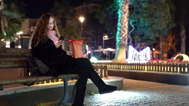Sonriente mujer relajada en ropa de invierno caliente sentado en el parque con decoraciones de Navidad por la noche y el uso de teléfono inteligente, mujer con pelo rizado navegar por Internet, chatear en línea y enviar — Vídeo de stock