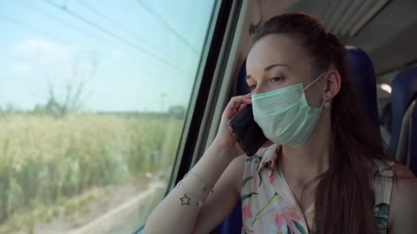 Woman in medical face mask driving by train, looking through the window, passing by railways stations and talking with friends by phone. Female passenger following all the rules and keeping social — 图库视频影像