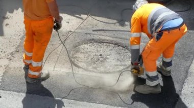 Hard working on construction site for opening clogged sewerage on the street, two road workers in protective uniform using heavy drilling electric hammer for breaking concrete on the sidewalk. Dust
