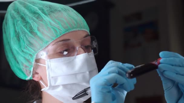 Female specialist in protective glasses, mask and gloves holding blood test tube in hands and writing down the analysis data on it, checking blood plasma of infected patients in laboratory and — Stok video