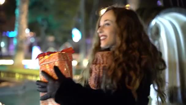 Medium shot of pretty smiling woman dancing in the park with christmas gift in hands on marble fountain background and street decorations, happy female enjoying winter holidays and traditions. Special — 图库视频影像