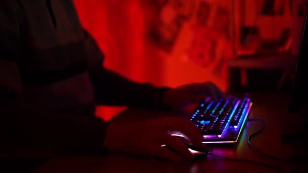 Gracz komputerowy siedzi przy stole w ciemnoczerwonym oświetlonym pokoju i trzyma turnieje online z wirtualnymi graczami w cyberprzestrzeni przy użyciu podświetlanej klawiatury widma i myszy. Online konkursy dla zabawy — Wideo stockowe