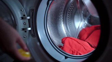 Çamaşır makinesini kirli çamaşırlarla dolduran kişi, içine şeffaf sarı kapsül atar ve makine kapısını kapatır. Ev hanımı, kirli çamaşırları yıkamaya hazırlıyor.