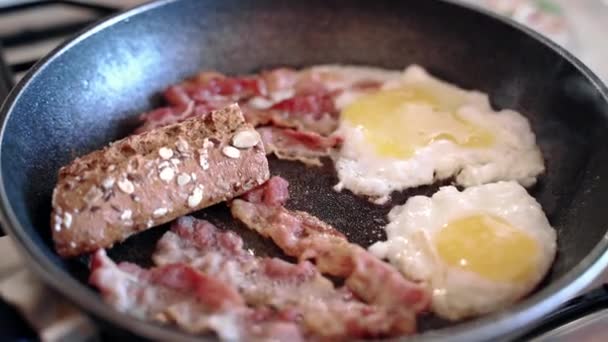Heerlijk Engels ontbijt op gasfornuis, knapperige spekreepjes, twee eieren en een schijfje roggebrood op een verwarmde pan met sissende olijfolie. Dagelijkse ontbijtbereiding, calorierijk — Stockvideo