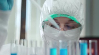 Koruyucu kıyafetli ve gözlüklü kadın laboratuvar araştırmacılarına steril ölçüm borusu kullanarak ve test tüplerinde çözelti bırakarak yakından bakın. Laboratuvarda tıbbi deneyler yapan konsantre bir kimyager.