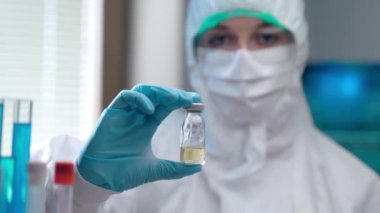 Koronavirüs enfeksiyonuna karşı potansiyel aşı tüpünün yakın plan çekimi koruyucu giyimli profesyonel kadın virologların elinde, kameraya bakan kendine güvenen bir laboratuvar çalışanı