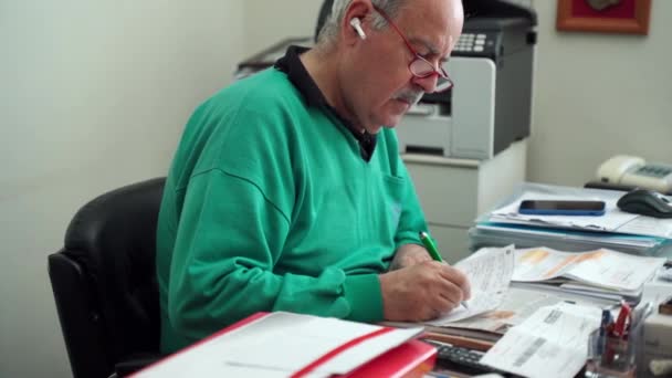 Trabalhador da agência idosa com fones de ouvido sem fio brancos sentados à mesa na sala de escritório e discutindo detalhes de compra ou venda de contrato de propriedade com os clientes. Acordo de leitura de homem, relatório de enchimento — Vídeo de Stock