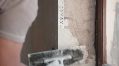Koruyucu eldivenlerle çalışan bir mühendis macun duvarları için metal spatula kullanıyor, spatulayı macunla dolduruyor ve beton duvar yüzeyini kaplıyor. İnşaatçı duvar ve açıyı düzleştiriyor