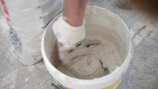 建筑工地上戴防护手套的工人使用小金属铲在塑料桶中搅拌石膏溶液。建筑工人为新公寓的油灰墙准备砂浆，水泥混合物 — 图库视频影像