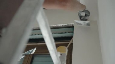 İnşaat alanındaki düz duvarları alçılayan merdiven tamircisinin düşük açılı görüntüsü. Duvar yüzeyini beyaz yapıştırıcı çözeltisiyle hizalamak için orta boy spatula kullanan usta bir müteahhit. Zor.