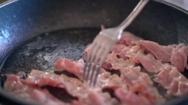 Sıcak tavada kavrulmuş taze pastırma şeritleri cızırdayan yağ, lezzetli sabah kahvaltısı için evde kızartılmış et dilimleri. Pastırma şeritlerini döndürmek için ahşap çatal kullanan kişi...
