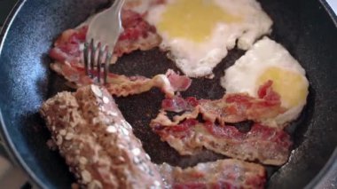 Sabah yağlı İngilizce ya da Amerikan kahvaltısı hazırlamak, evde ısıtılmış tavada sağlıksız yemek pişirmek. Kızarmış pastırma dilimleri ve yumurta için çatal kullanan biri.