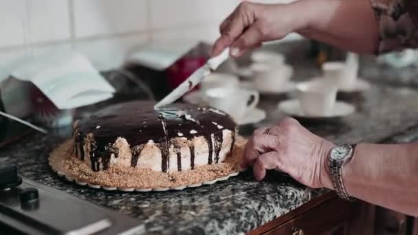 Manos de mujer mayor sosteniendo cuchillo de cocina y cortando delicioso pastel de chocolate en la mesa de la cocina sobre fondo borroso de tazas de café. Primer plano de sabroso pastel dulce calórico cortado en muchas piezas para — Vídeo de stock