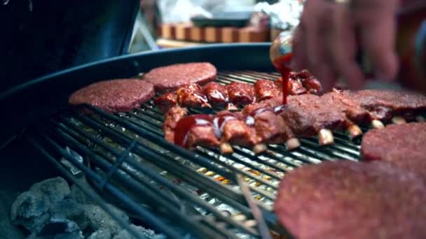 Chef derramando molho picante artesanal em costelas de porco assadas e hambúrgueres fritando em grelhas ao ar livre, molho especial para churrasco com ervas aromáticas e especiarias, tempero de carne assada para piquenique de churrasco — Vídeo de Stock