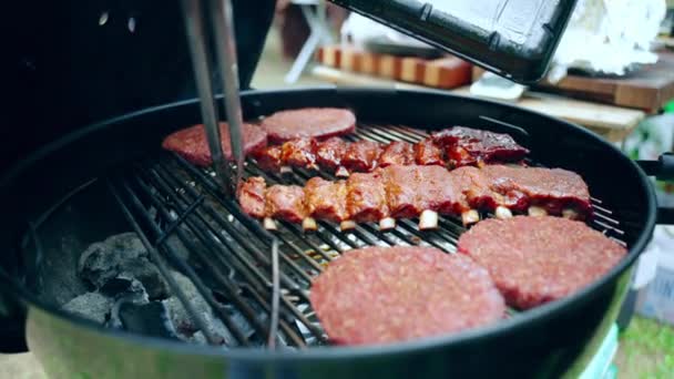 Fin de semana de relax americano con deliciosa parrilla de barbacoa al aire libre, hombre asando costillas de cerdo y hamburguesas en parrilla de barbacoa, comprobando la preparación de la carne con pinzas de cocina y vertiendo salsa aromática picante a base de hierbas — Vídeos de Stock