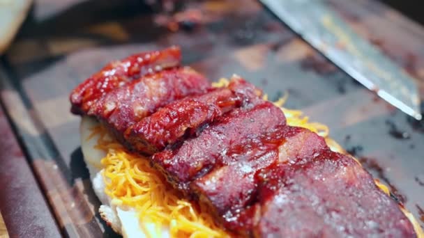 Gustoso hamburger preparato su tavolo da cucina in legno con coltello professionale disteso nelle vicinanze, panino arrosto farcito con fette di carota piccanti e costolette di maiale succose alla griglia. Delizioso ma malsano cibo di strada — Video Stock