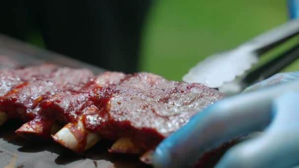 Festeggiando la festa barbecue all'aperto, chef in guanti di gomma gettando le ossa dalle costole di maiale arrosto con pinze metalliche. Primo piano di succose costolette di carne affumicata sul tagliere di legno, delizioso — Video Stock