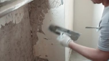 Pencerenin altında metal mala ve beyaz alçı macunu kullanarak duvarları spatula ve macun kaplamalarıyla düzleştiren iş kıyafetleriyle çalışan tamirci. Duvarların yenilenmesi