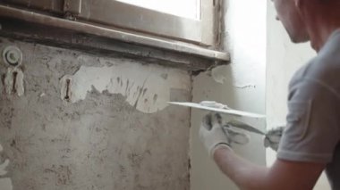 İş kıyafetleri içinde usta bir tamirci. Metal mala, spatula ve beyaz macun kullanarak pencerenin altına eski püsküllü beton duvarları kokutuyor. Duvarın yüzeyini macunla kaplıyor. Duvarlar