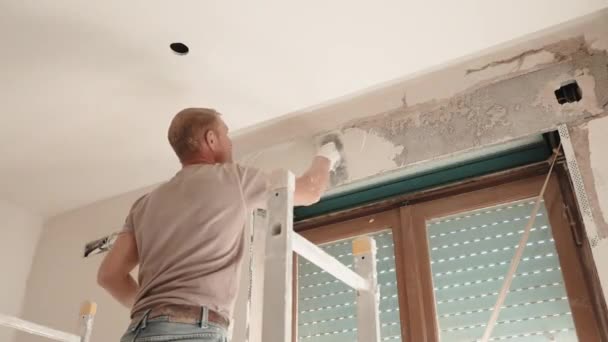 Låg vinkel syn på professionell byggarbetare inriktning och utjämning putsade väggar med vit efterbehandling pasta lösning ovanför fönstret med gamla externa rullgardiner. Byggare i skydd — Stockvideo