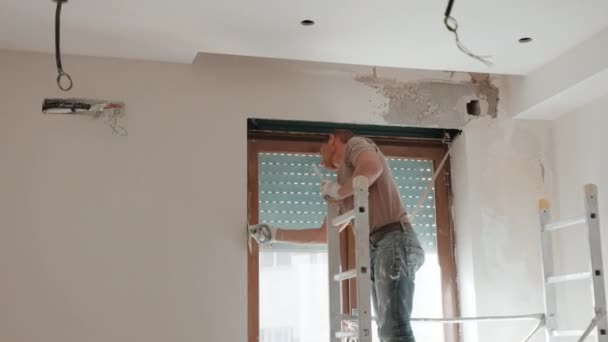 Travaux de réparation sur chantier dans un nouvel appartement, maître ingénieur debout sur l'échelle et mur écartant autour de la fenêtre avec stores à rouleaux externes. Travailleur utilisant une spatule métallique pour aligner — Video