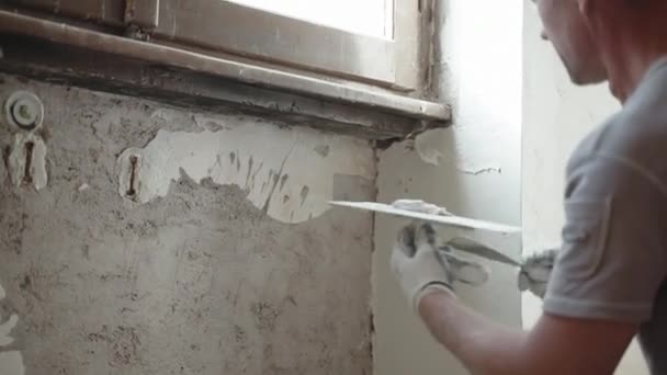 Réparateur qualifié en vêtements de travail en plaçant un vieux mur de ciment déchiqueté sous la fenêtre à l'aide d'une truelle métallique, d'une spatule et d'une pâte à tartiner blanche, d'une surface murale nivelée par l'homme avec du stuc de mastic. Murs — Video