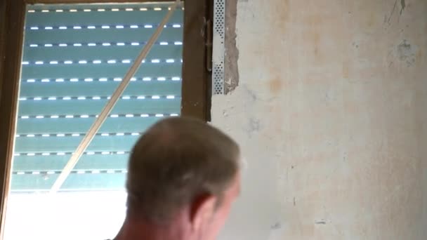 Constructor masculino que hace la reconstrucción de ángulos de pared, reparador usando espátula para alinear y alisar ángulos metálicos. Vista trasera de la pared salpicada de trabajadores cerca de la ventana con persianas rotas externas — Vídeo de stock