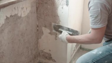 İş kıyafetleri içinde usta bir tamirci pencerenin altında metal mala ve beyaz alçı macunu kullanarak eski yırtık beton duvarları alçılıyor, duvar yüzeyini spatula ve macun kaplamasıyla düzleştiriyor. Duvarlar