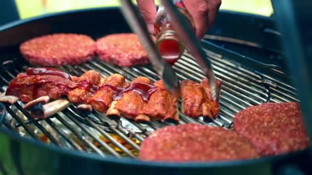 Homem derramando molho de tomate em costelas de porco prontas e óleo com escova de cozinha de silicone. Processo de preparação de churrasco de carne na natureza, hambúrgueres suculentos assando na grade de churrasco na queima de carvão vegetal. Fim de semana — Vídeo de Stock