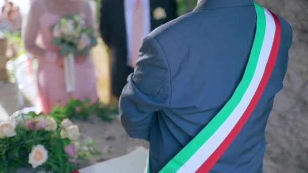 Cerimônia de casamento tradicional italiana ao ar livre sob o arco antigo decorado com flores frescas perto do restaurante, oficiante de casamento com fita de bandeira em todo o ombro segurando promessas de casamento — Vídeo de Stock