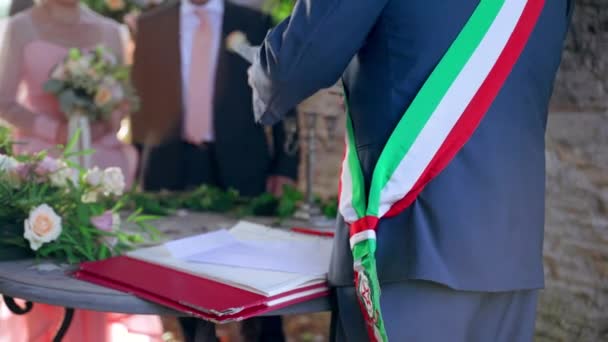 Ceremonia de boda italiana tradicional al aire libre bajo el antiguo arco decorado con flores frescas, vista trasera del oficiante de la boda con cinta de la bandera a través del hombro preparando el certificado de boda para — Vídeo de stock
