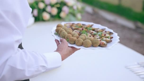 Pincér egyenruhás tányérokban sült olajbogyóval és tenger gyümölcseivel és bankettasztal esküvői partin, szolga ételmenüt készít a meghívott vendégeknek. Tengeri ételek választéka a büfében — Stock videók