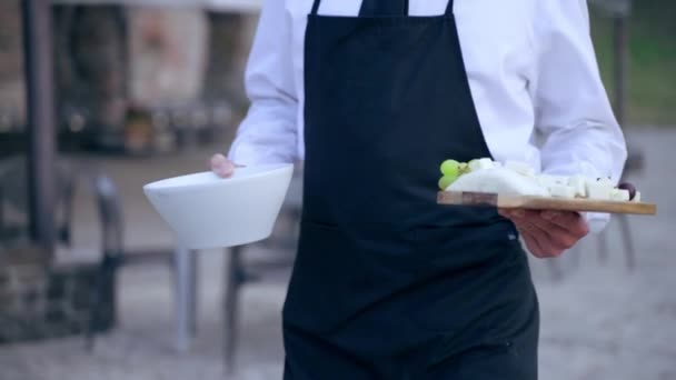 Garçom em uniforme andando até a mesa do banquete em câmera lenta segurando em mãos tábua de madeira com queijo picado, mel e uvas, servo de restaurante de alto nível preparando mesa buffet de enchimento com — Vídeo de Stock