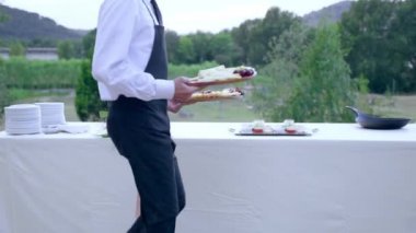 Luxury catering food service in elegant restaurant with banquet table arranged outdoors, waiter in uniform holding wooden trays with chopped cheese and grapes. Tasty traditional appetizers and snacks