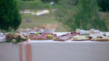 Dolly shot of appetizing banquet table outdoors decorated with fresh flowers bouquets on background of amazing meadow and lake. Luxury buffet table full of snacks and canapes, fresh tasty meat slices