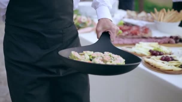 Профессиональный официант в форме кладет черную сковородку с закусками на банкетный стол на роскошной свадебной вечеринке на открытом воздухе на ресепшене ресторана, слуга накрывает стол для приглашенных гостей с разнообразием — стоковое видео
