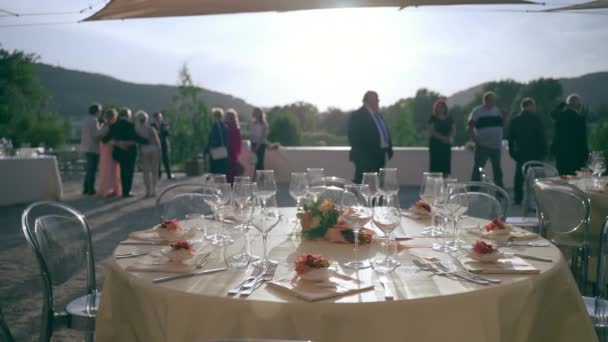 Des invités de mariage flous attendent le couple marié au premier plan de la décoration des tables de fête servies. Vous pourrez prendre des photos au coucher du soleil, parler et vous détendre devant le restaurant de luxe. Service traiteur et — Video