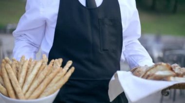 Waiter in uniform walking to the banquet table in slow motion holding in hands trays with bread slices and crispy breadsticks, high level restaurant servant preparing buffet table filling with snacks