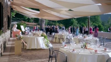 Blurred wedding guests waiting for married couple on foreground of served festive tables decor. Beautiful round tables decorated with flowers bouquets and elegant tableware and cutlery. Wedding party